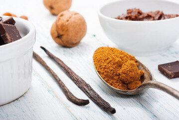 pile of brown sugar on old spoon, vanilla, broken dark chocolate blocks on white wooden table