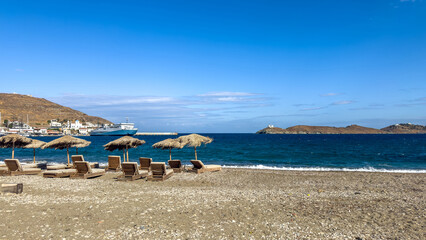 Fototapeta premium Peaceful sunny beach featuring straw umbrellas and wooden loungers facing the deep blue Aegean Sea within Coressia, Kea (Tzia), Greece, with the ferry docked near the harbor under a clear sky