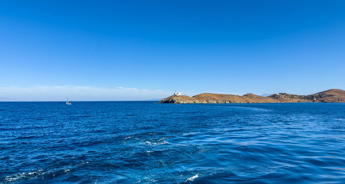 Bright midday view of the Kea (Tzia) island coastline featuring the small lighthouse upon a rocky cape, deep blue Aegean Sea waters, and a distant sailboat under a clear summer sky