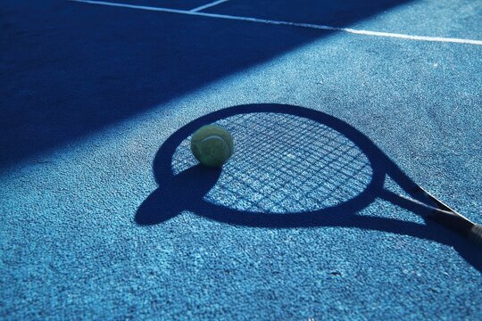 Shadow of a tennis racket and ball cast on a blue court during a sunny afternoon, illustrating the sport's dynamic nature and playfulness in a vibrant setting
