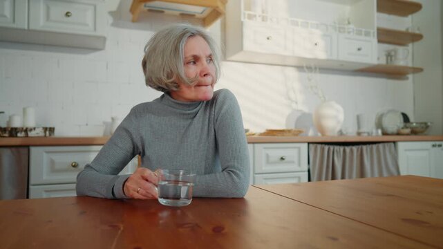 Old woman drinking water in modern home kitchen, portrait of aged lady indoors. Worried elderly widow sitting alone and remembering life, sad grey-haired female person thinking about past, melancholy