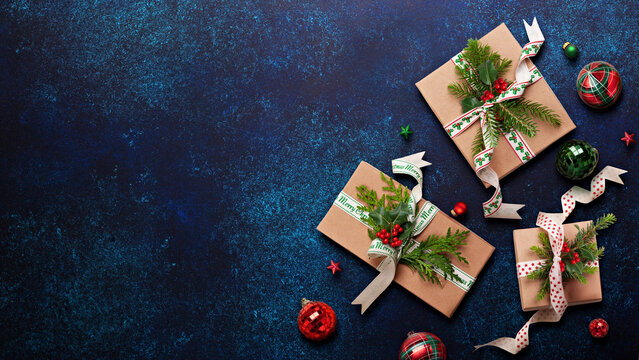 Top view of wrapped Christmas gifts with natural decor and ornaments scattered on a rich blue background.