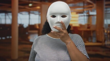 Woman holds white mask in restaurant building with one hand pressed against its surface amid empty tables and columns; mystery anonymity. - Powered by Adobe
