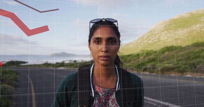 Standing Indian woman wearing bomber jacket on paved coastal road, with backpack and sunglasses