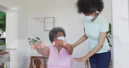 Senior woman wearing mask holding pink dumbbells as caregiver wearing polo guiding movement at home