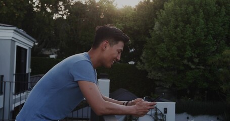 Leaning Asian man checking smartphone on balcony of modern house with metal railing and big windows