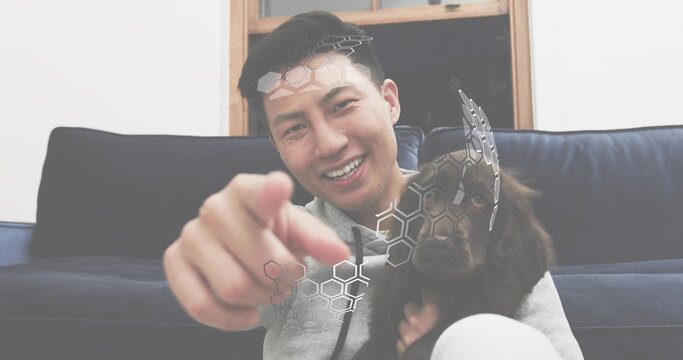 Smiling man holding dog on living room couch under window, pointing at camera with hexagon overlay