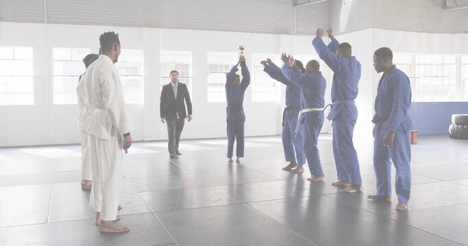 Holding gold trophy overhead, blue-gi student celebrating in training hall on mats with tire stacks