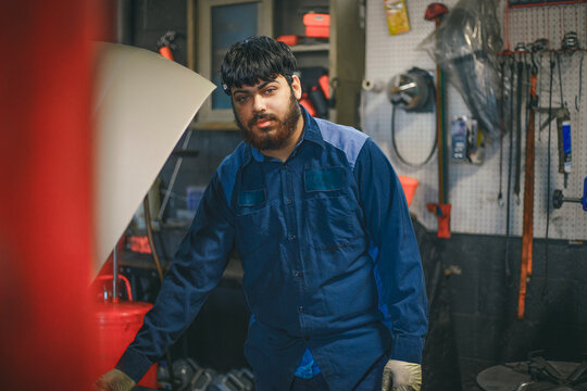 A mechanic stands near a car in a workshop, wearing a blue uniform.