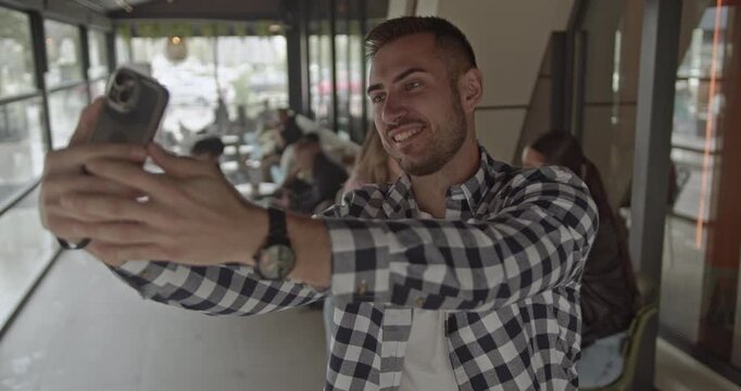 Young man takes a selfie with his smartphone, smiling and posing. The people in the background are out of focus, suggesting a bustling environment. Capturing fun moments and sharing online.