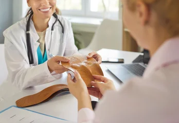 Selbstklebende Fototapeten Restaurant Smiling doctor showing a pair of orthopedic insoles to woman patient during medical consultation in clinic. Foot posture correction, treatment of flat feet. Orthopaedics, healthcare concept  © Studio Romantic