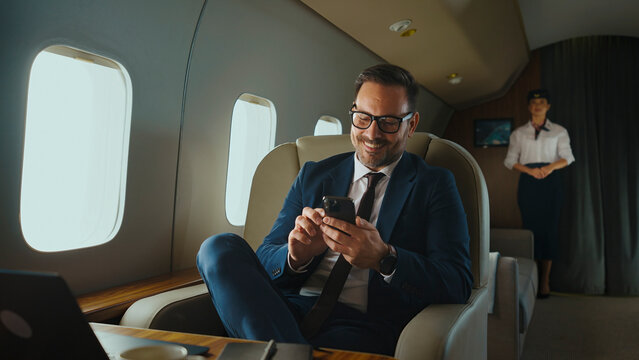 Business executive in a tailored suit and glasses smiles while using a smartphone in a luxurious private jet cabin, flight attendant in the background symbolizing business travel and connectivity - Powered by Adobe