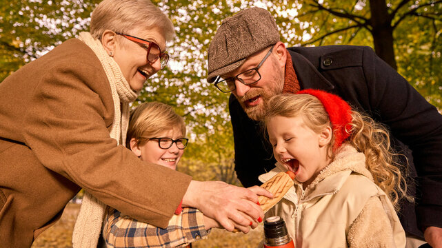 Grandmother and father helping girl eat hotdog during autumn picnic. Concept of care, education, and intergenerational harmony for food branding and lifestyle storytelling.