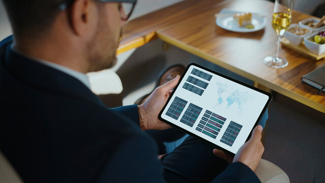 Businessman holding a digital tablet with financial charts and global market data, reviewing analytics in a sophisticated environment with a glass of champagne and snacks on a private jet
