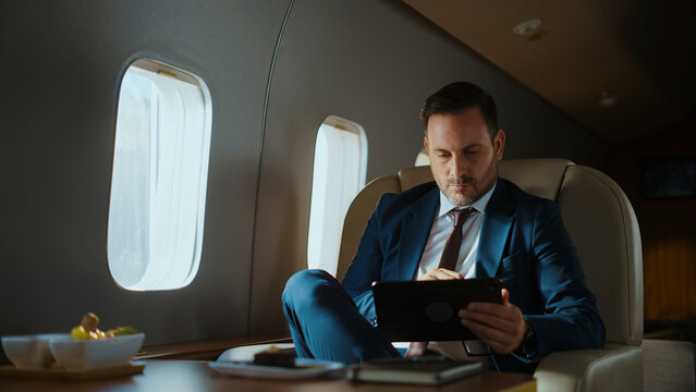 Businessman in a suit focusing on his digital tablet while relaxing on a comfortable seat inside a luxurious private jet cabin, enjoying premium corporate travel