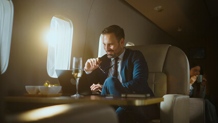 Businessman in blue suit smiles confidently on a private jet, working on a laptop with champagne at...