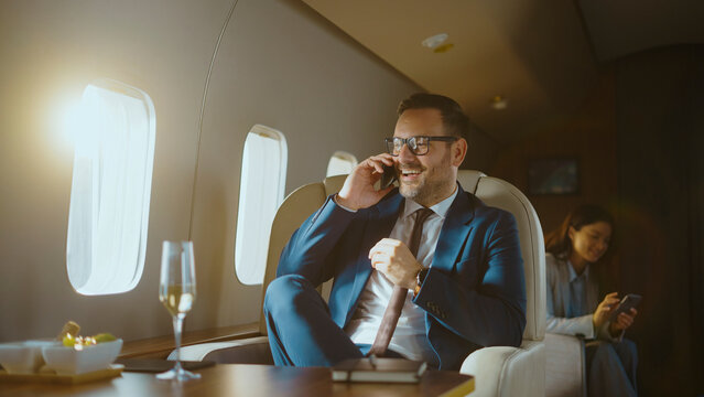 Successful businessman smiling and talking on a smartphone, enjoying a luxury corporate flight in a private jet, with a colleague using her phone in the background