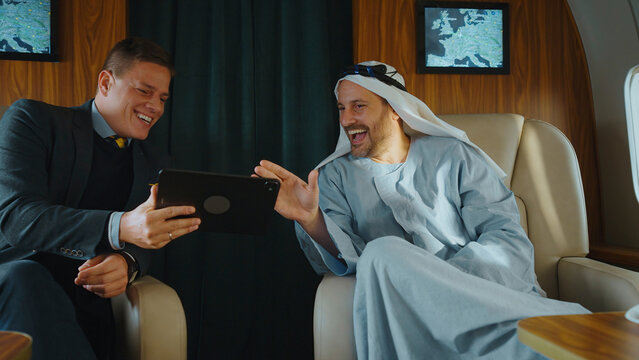Two diverse businessmen laughing and discussing a deal while traveling in a luxurious private jet, sharing a digital tablet and enjoying a moment of successful collaboration