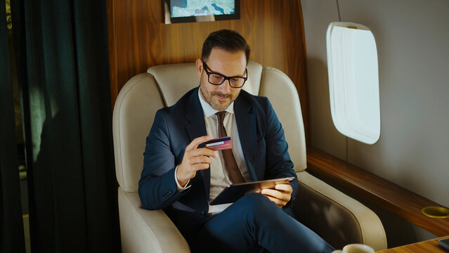 Man in a business suit using a credit card and digital tablet for online shopping or a financial transaction while traveling comfortably in a luxurious private airplane