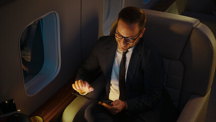 Smiling businessman flying in a private jet cabin during a night flight, using a digital tablet for work, showcasing luxury business travel and connectivity concepts