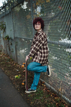 Teen with curly hair in plaid shirt holding skateboard by fence outdoors