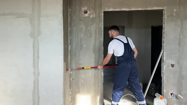 A bearded man in a work uniform is priming the walls after applying gypsum plaster. 