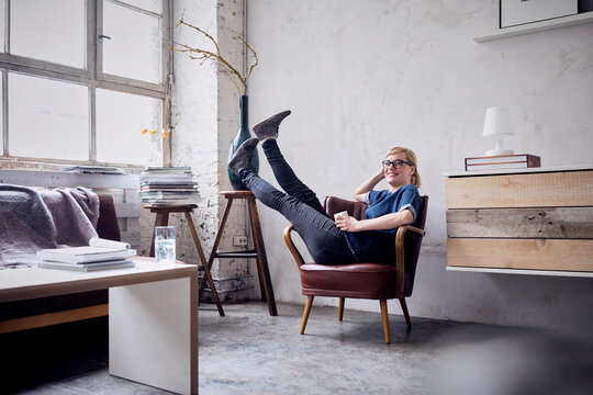 Happy woman sitting with legs in the air on armchair in loft