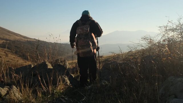 A lonely male tourist with a backpack and a walking stick walks along a rocky mountain trail at sunset. Traveling to unexplored corners of the world. Solo mountain hiking adventure.