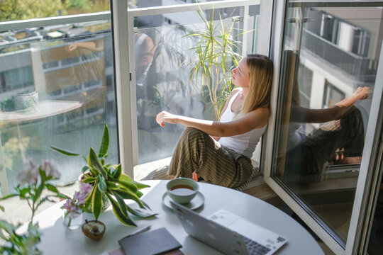 Young woman relaxes during remote work by window with laptop and coffee