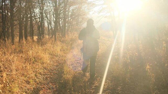 A lonely traveler walks along a forest road illuminated by the rays of the sunrise or sunset, carrying a large heavy backpack. The man travels alone without anyone else. Solo forest journey.