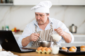 Man in chef hat and apron prepares cupcakes in kitchen according to recipe from the Internet - looking at laptop screen