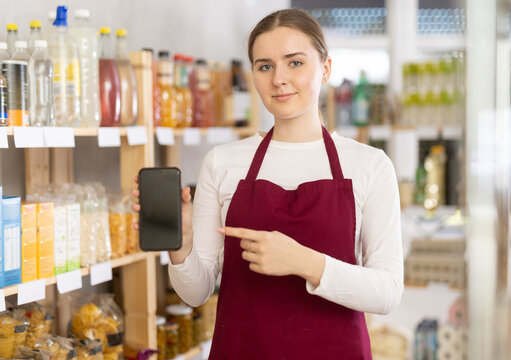 Girl seller in shop hold out hand with phone, points at screen with finger