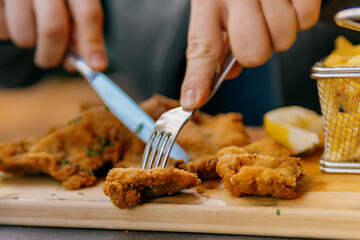 Close-up of a person cutting a piece of crispy, golden-brown fried schnitzel served on a wooden board with a side of french fries and lemon.