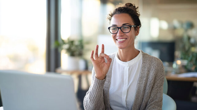 Deaf businesswoman using sign language while smiling at her laptop in a modern office, showcasing professional communication and inclusivity - Powered by Adobe