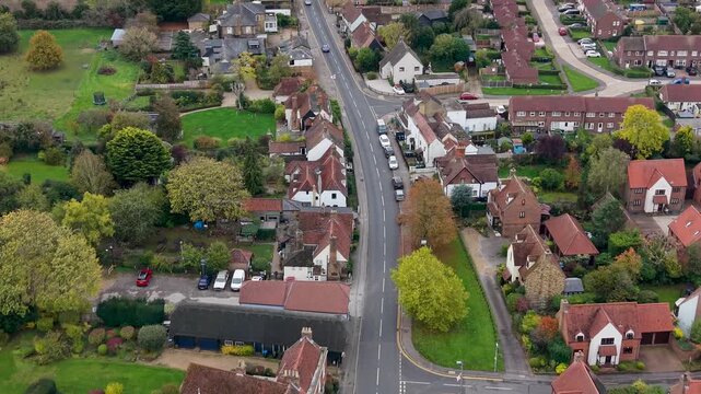 Drone flies forward up a hill through Roydon, Essex, along a quiet village road lined with houses and shops. Cars pass as the drone follows the street to a T junction, showing trees and grass nearby.