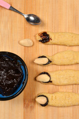 An overhead shot showing the decoration of baked witch finger cookies by applying dark plum jam with a spoon to attach the almond nails
