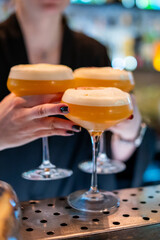 A bartender's hands hold three Passion Star Martinis, a popular cocktail with a vibrant orange hue, topped with white foam, ready to be served on a bar counter