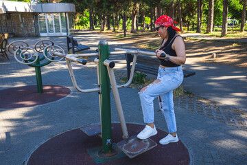 Fototapeta premium A woman exercises on outdoor fitness equipment in a sunny park surrounded by trees. The setting shows an urban recreational area with benches and fitness machines under natural daylight.