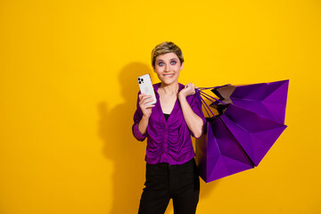 Young stylish woman smiles as she selfies with a smartphone and purple shopping bags against a bright yellow backdrop
