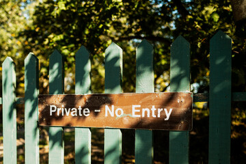 Home made Private - No Entry sign seen in a painted wooden gate at the entrance to a woodland home in rural Britain. Part of the overgrown garden lies beyond the painted gate.
