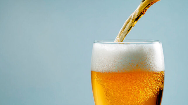 Cold golden beer being poured into a glass with thick foam head against a light blue background