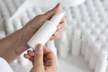 A worker's hands hold a blank white pump bottle for inspection before labeling. The background shows rows of blurred, identical blank cosmetic bottles in this organic production lab.