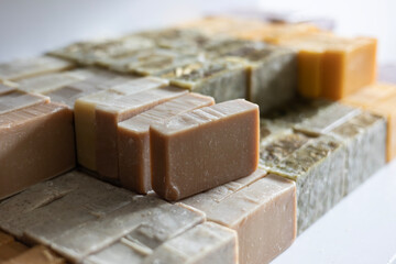 Various bars of handmade, cold-process soap are curing on a shelf. These organic cosmetics feature different natural ingredients, resulting in beige, green, yellow, and purple colors.