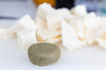 A round, textured solid shampoo bar sits on a white surface. In the blurred background are large chunks of raw shea butter, a key ingredient for making natural and organic cosmetics.