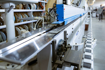 A close-up view of an automated edge banding machine in a furniture factory. The conveyor chain and metal guides are visible, with shelves of edge band spools in the background.