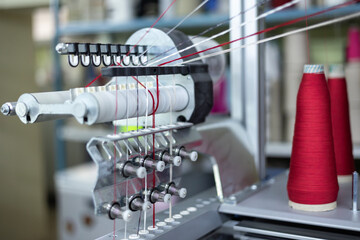 A close-up view of an industrial knitting machine in a knitwear factory. Red and white threads are fed from large cones through the guides and tensioners of the automated equipment.