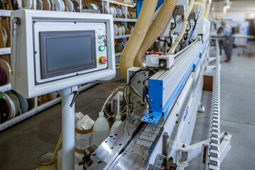 A close-up of the edge banding machine's control panel. The touchscreen HMI operates the automated system in a modern furniture factory, with the conveyor and shelving in the background.