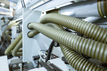 A close-up of an automated edge banding machine in a furniture factory. Corrugated plastic hoses for dust extraction are connected to the machine, which glues and cuts edges on MDF and chipboard.