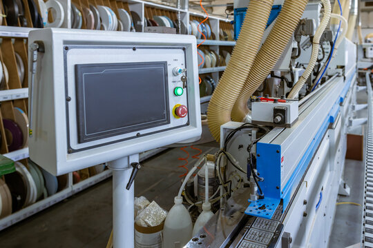 A close-up of the edge banding machine's control panel. The touchscreen HMI operates the automated system in a modern furniture factory, with the conveyor and shelving in the background.
