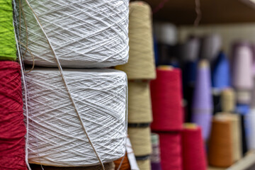 A close-up, selective focus view of large white yarn cones stacked on a shelf in a knitwear factory. Colorful bobbins of red, green, and beige thread are blurred in the background.